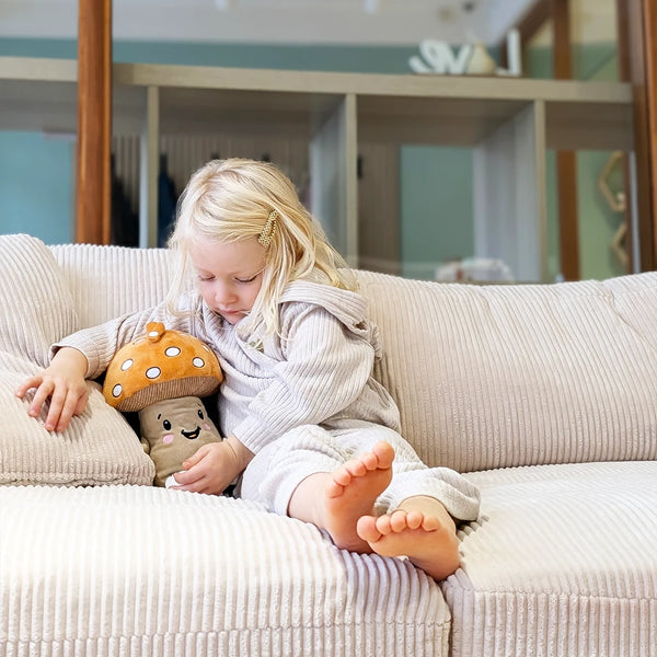 Kleines Mädchen sitzt gemütlich auf dem Sofa und kuschelt mit Lenabie Felix (Pilz) – das weiche Audio-Kuscheltier als beruhigender Begleiter für entspannte Momente zu Hause.
