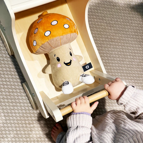 Lenabie Felix (Pilz) sitzt im Spielzeug-Puppenwagen, während ein Kind ihn schiebt – das weiche Audio-Kuscheltier als fröhlicher Begleiter im Alltag und beim Spielen.