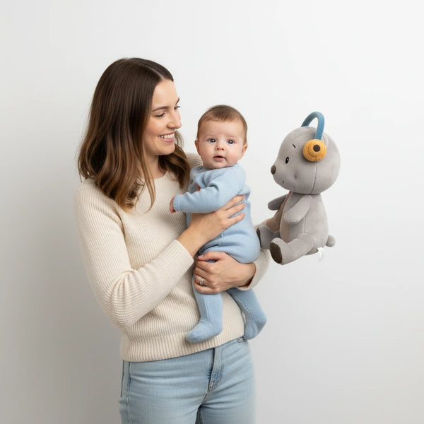 Mutter hält ihr Baby im Arm, daneben das Lenabie Aaron (Bär) mit Kopfhörern – emotionales Produktbild zum Lenabo Starter Kit mit Lenabo Smart Speaker, zeigt das smarte Spielsystem ab Geburt (0+), das Geborgenheit, Audioinhalte und frühkindliche Förderung vereint.